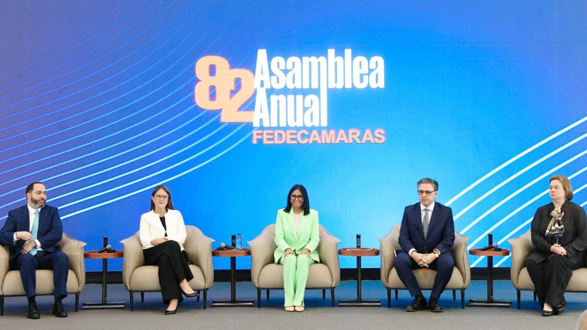 Delcy Rodríguez, presidenta encargada de Venezuela, sentada al centro del escenario de la 82.ª Asamblea Anual de Fedecámaras, junto a otros cuatro asistentes, frente a una pantalla LED azul con el nombre del evento